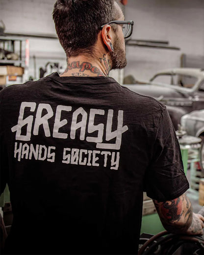 Person wearing a black t-shirt with 'Greasy Hands Society' text in a workshop setting - Greasy Hands Society Men's logo graphic tee