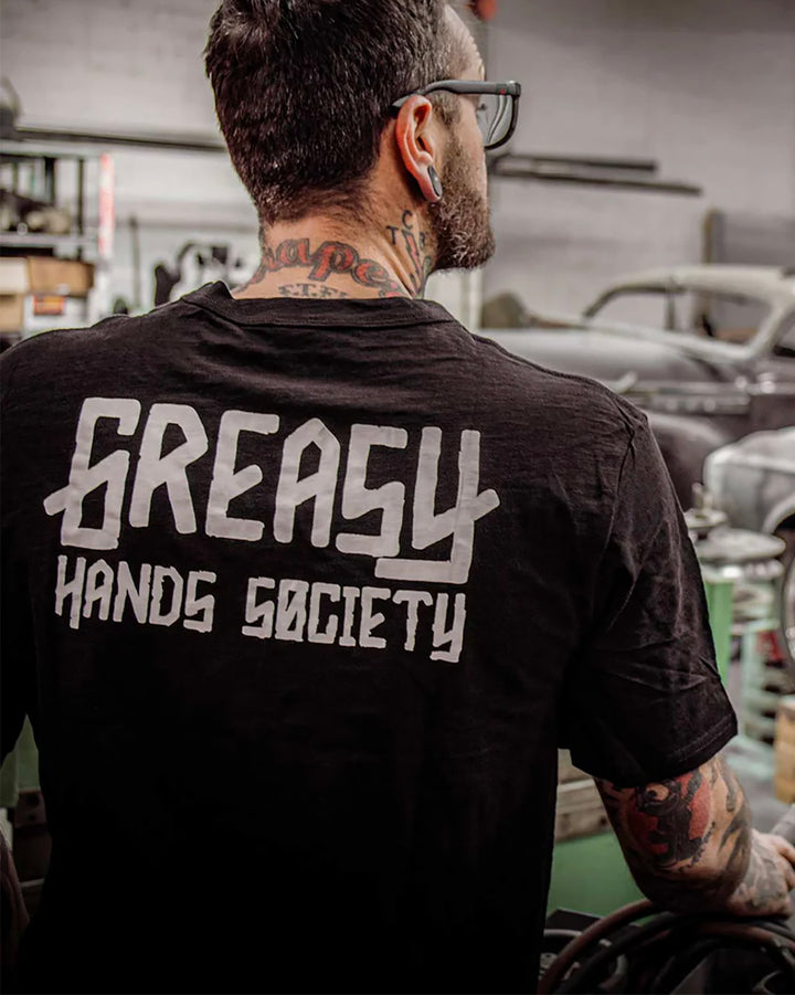 Person wearing a black t-shirt with 'Greasy Hands Society' text in a workshop setting - Greasy Hands Society Men's logo graphic tee