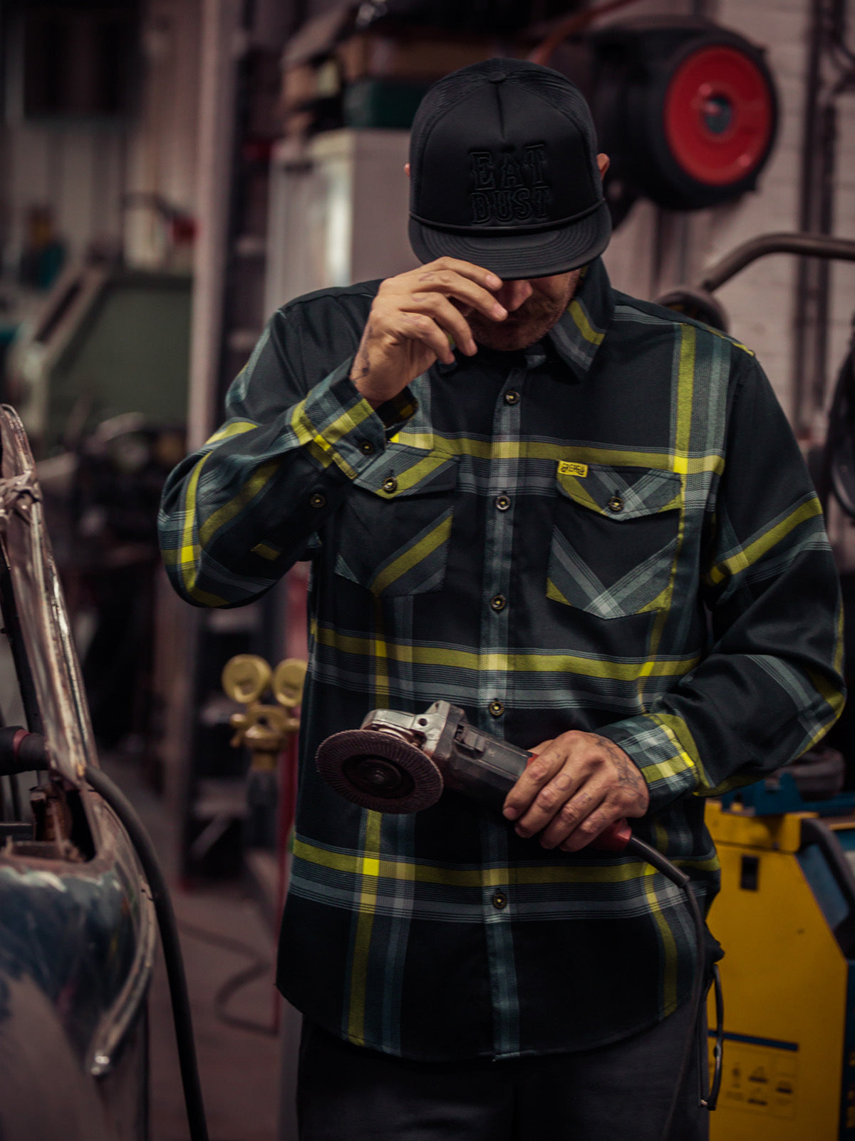 Person wearing a black and yellow plaid shirt and cap, holding a tool in a workshop setting - Eat Dust Flannel by Greasy Hands Society