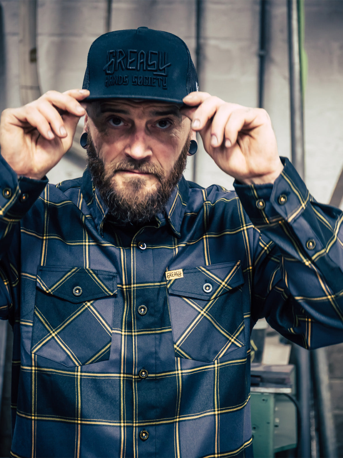 Man wearing a black gray and orange plaid flannel shirt and cap with 'Greasy Hands Society' logo, standing indoors - El Lobo Flannel by Greasy Hands Society