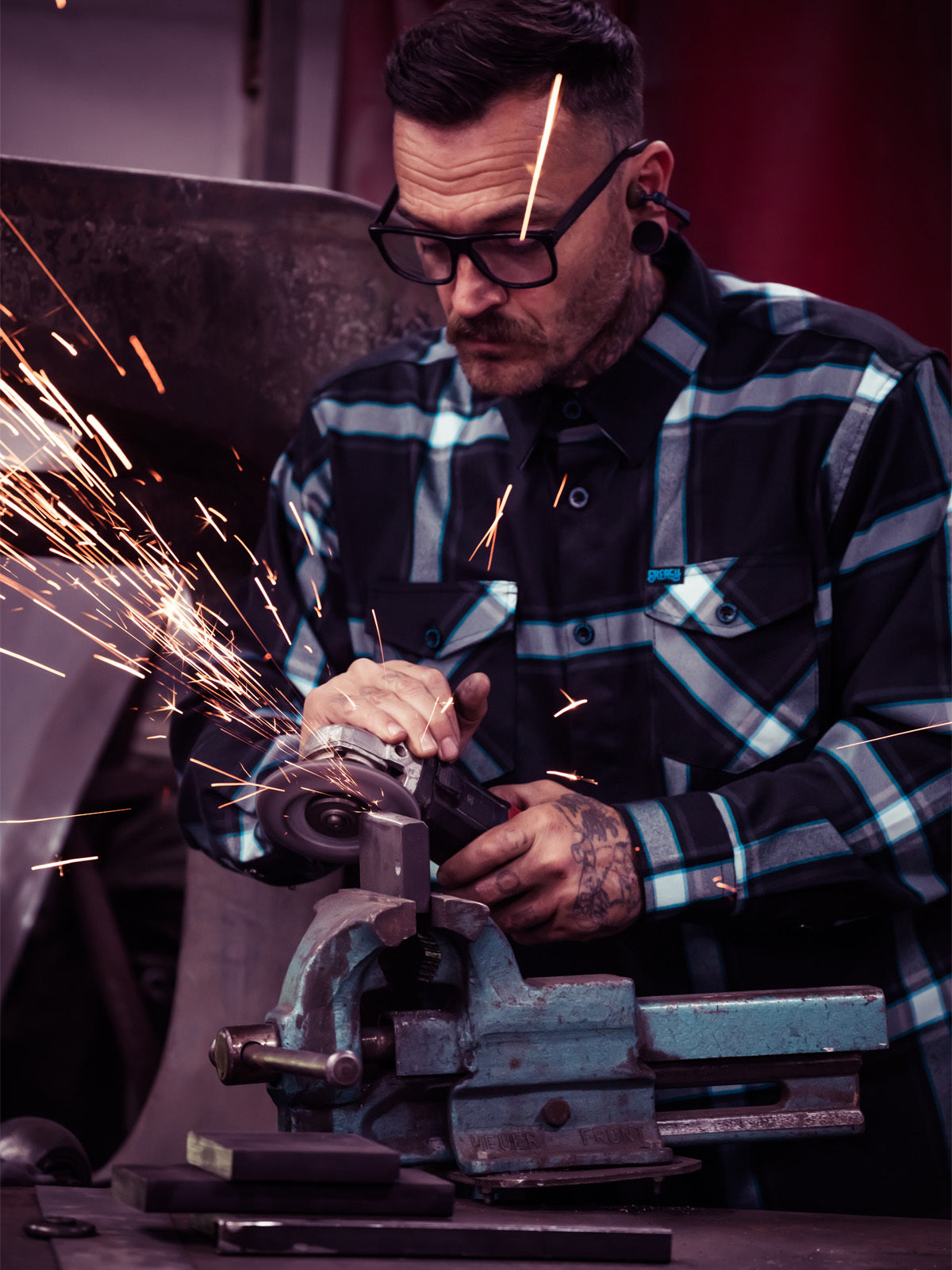 Man welding metal with sparks flying, wearing a blue and black plaid flannel shirt - Gravity Fed Flannel by Greasy Hands Society