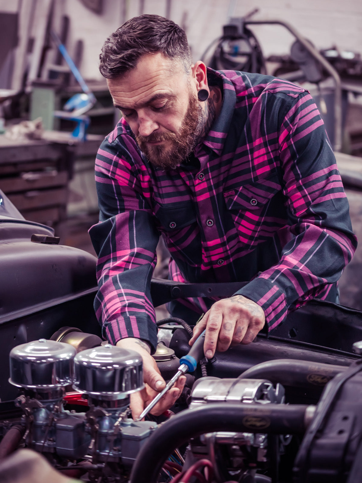 Man in a pink and black plaid flannel shirt working on a car engine in a garage - Lonely Heart Flannel by Greasy Hands Society
