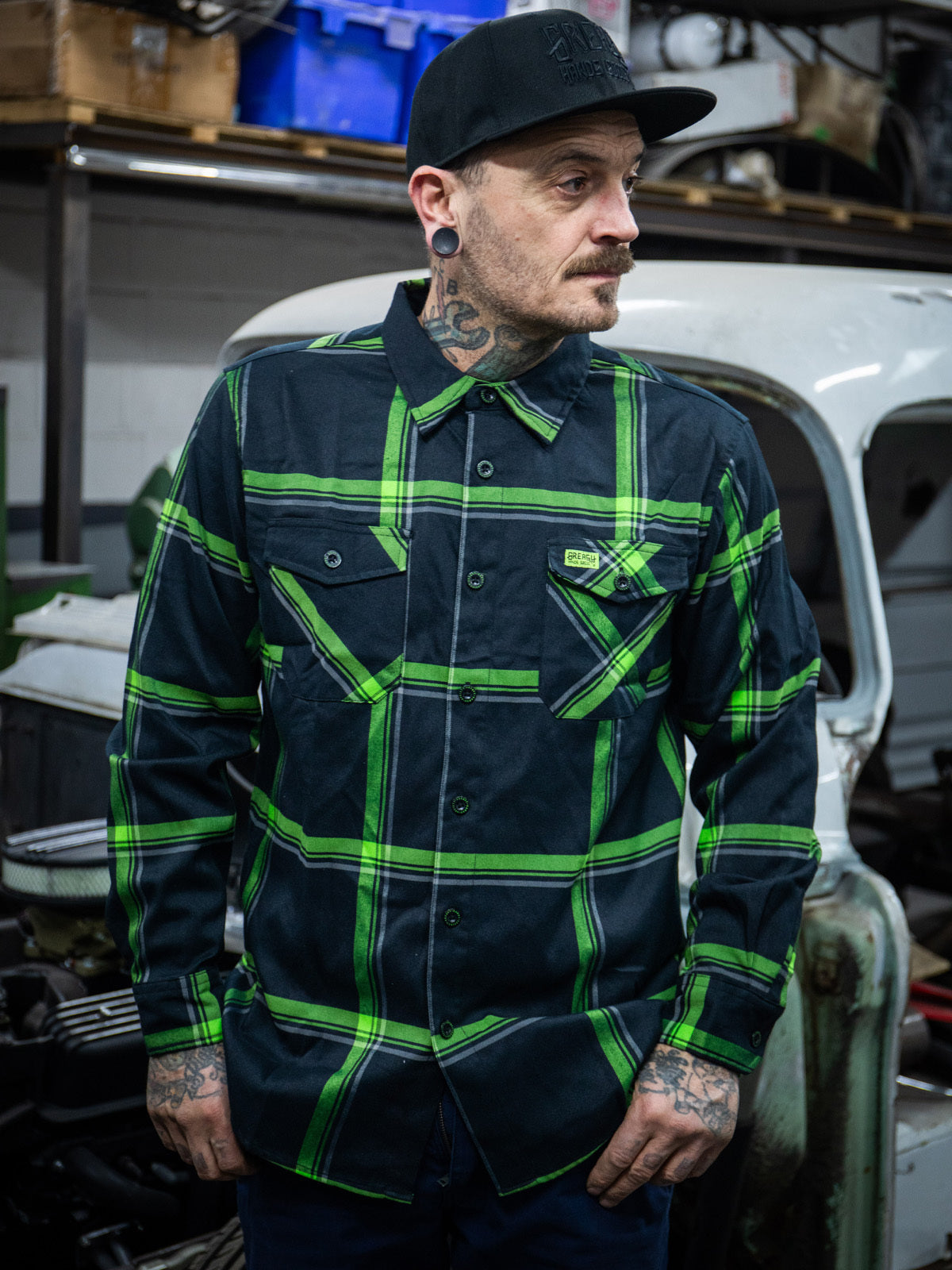Man wearing a black and green plaid shirt in a workshop setting - The Moto Flannel by Greasy Hands Society