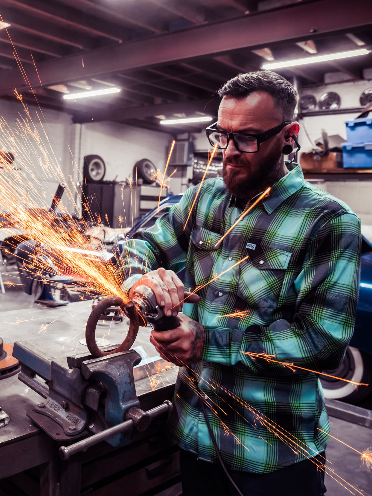 Man wearing. a blue and green flannel shirt using a grinder with sparks in a workshop setting - The Sucker Punch Flannel by Greasy Hands Society