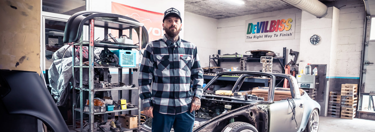 Man standing in a garage with automotive parts and a car in the background - The Banger Flannel by Greasy Hands Society