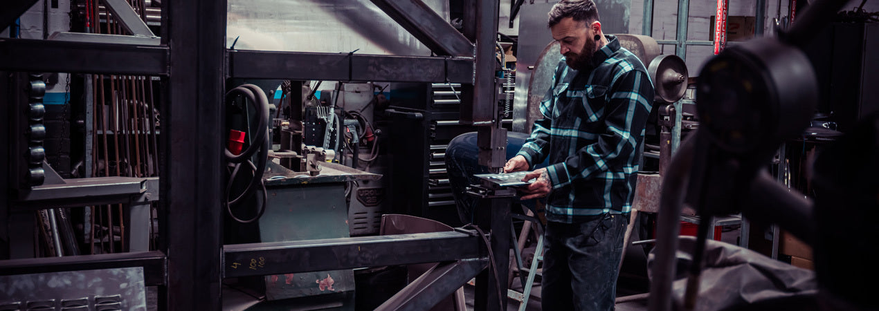Person working in a workshop wearing a blue and black plaid flannel shirt with tools and equipment around - Gravity Fed Flannel by Greasy Hands Society