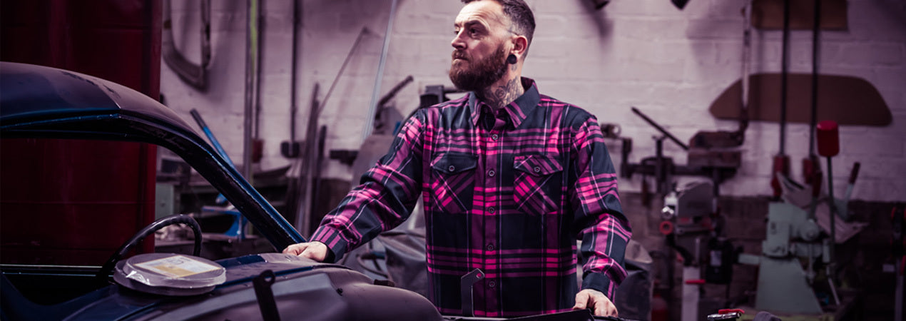 Man in a pink and black plaid shirt standing next to a car in a workshop - Lonely Heart Flannel by Greasy Hands Society