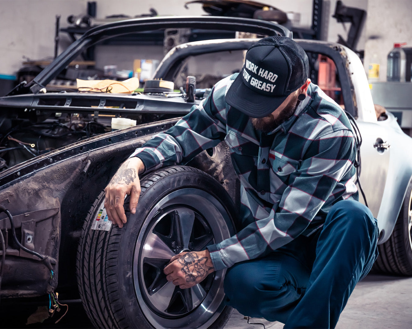 Person working on a car tire in a garage wearing a cap with text - The Banger Flannel by Greasy Hands Society
