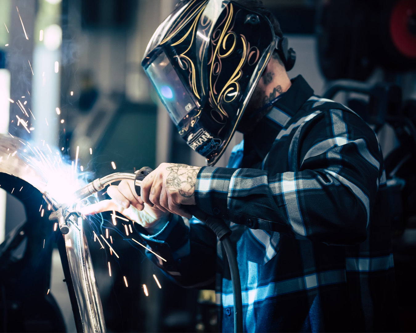 Person in a black and blue plaid shirt welding with protective gear in a workshop setting - Gravity Fed Flannel by Greasy Hands Society