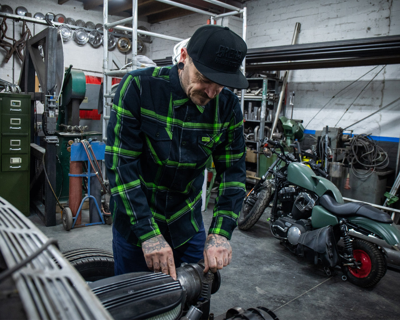 Man working on a classic car wearing a green and black flannel shirt in a workshop with tools and equipment around - The Moto Flannel by Greasy Hands Society