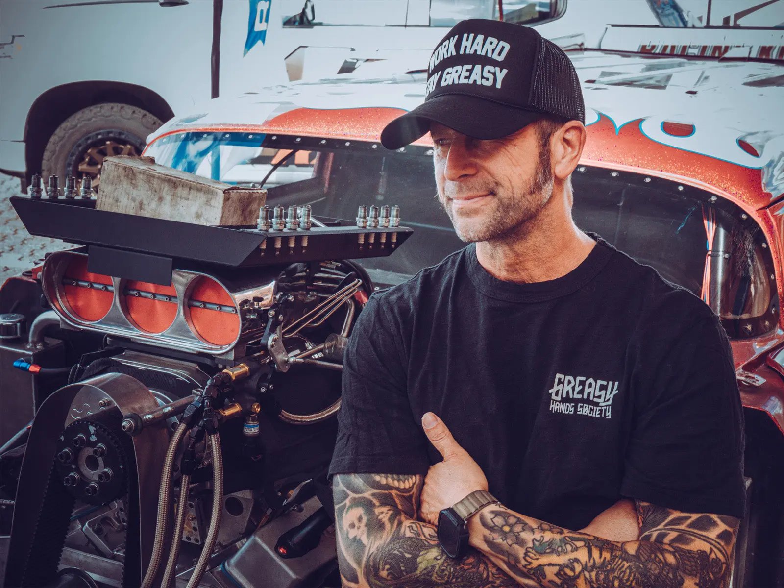 Man wearing a black cap and t-shirt with a tattooed arm standing in front of a race car - the greasy hands society logo tee by greasy hands society