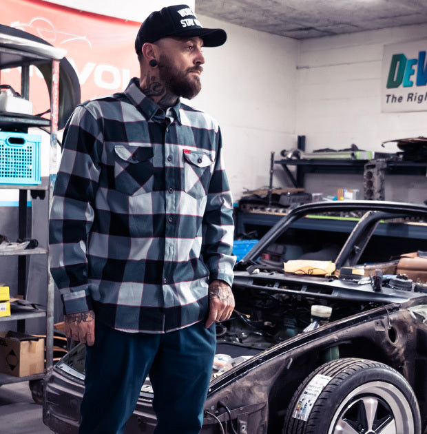Man in a checkered black and gray shirt and cap standing next to a car in a garage - The Banger Flannel by Greasy Hands Society
