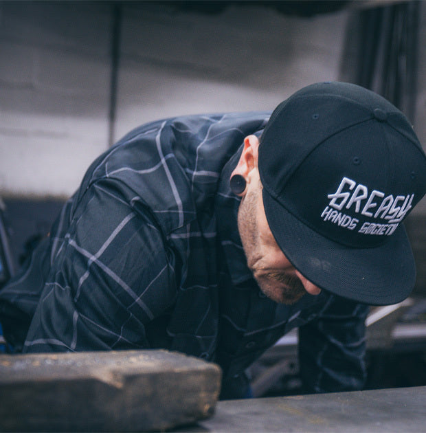 Person wearing a cap with 'Greasy Hands Society' text, and a gray plaid flannel shirt working on a tire - The Dapper Flannel by Greasy Hands Society
