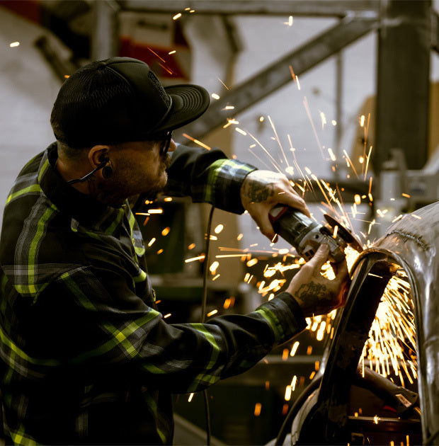 Person wearing a black and yellow plaid flannel shirt welding metal with sparks flying in a workshop setting - Eat Dust Flannel by Greasy Hands Society