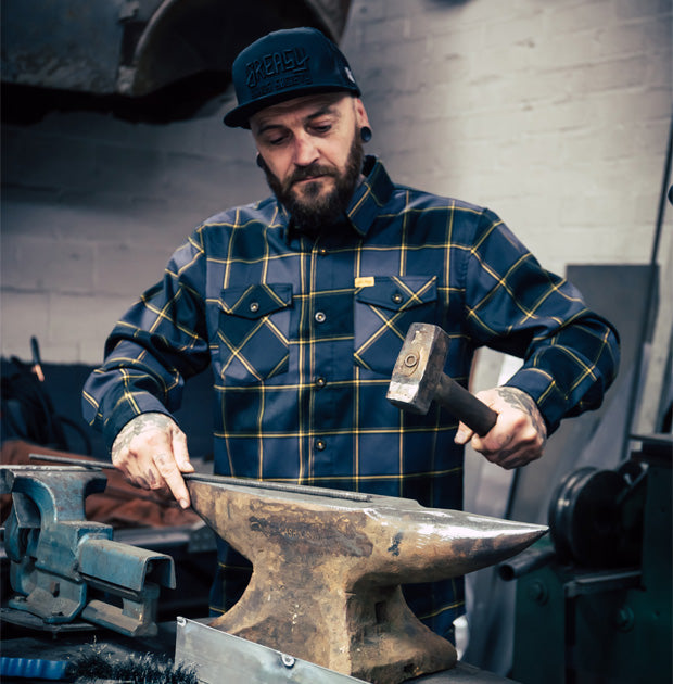 Man wearing a gray orange and black plaid flannel shirt working with a hammer on an anvil in a workshop setting - El Lobo Flannel by Greasy Hands Society