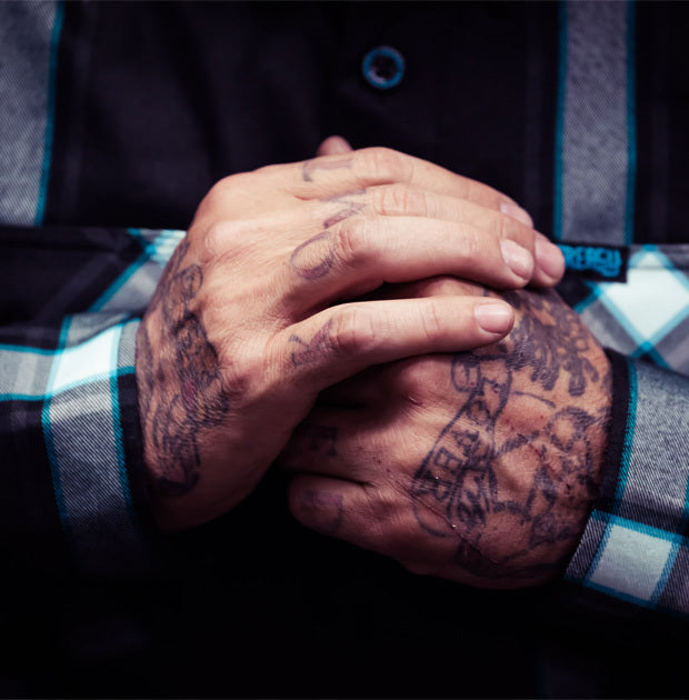 Close-up of tattooed hands clasped together with a black and blue plaid shirt background - Gravity Fed Flannel by Greasy Hands Society