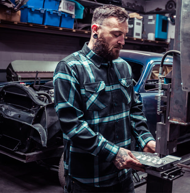 Man in a black and blue plaid shirt working on a car in a garage - Gravity Fed Flannel by Greasy Hands Society