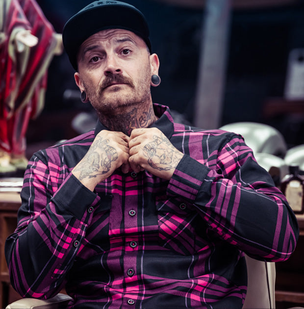 Man wearing a black and pink plaid shirt with tattoos on his arms, sitting in a room - Lonely Heart Flannel by Greasy Hands Society