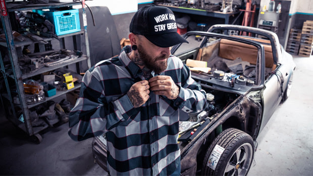 Man in a black and gray checkered shirt with red line and cap in a workshop with tools and equipment around - The Banger Flannel by Greasy Hands Society