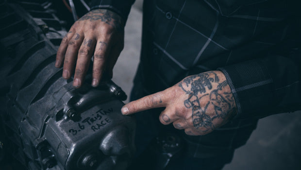 Person pointing to a part of an engine with visible tattoos on their hands - The Dapper Flannel by Greasy Hands Society