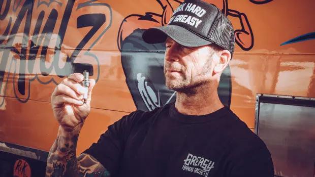 Man holding a piece of a metal screw in front of a graffiti-style wall wearing a black shirt and lack hat - the greasy hands society logo tee by greasy hands society