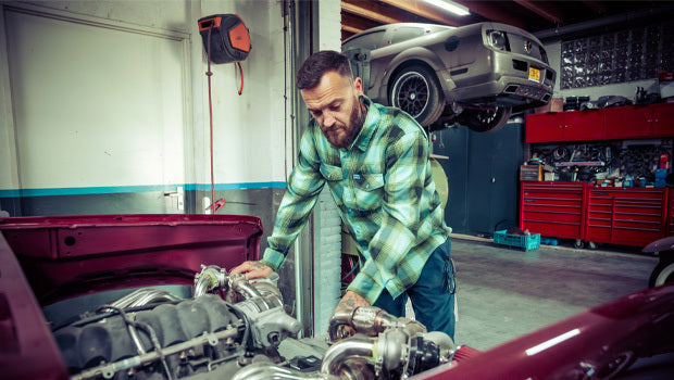 Man working on a car engine in a garage in a blue and green flannel - The Sucker Punch Flannel by Greasy Hands Society