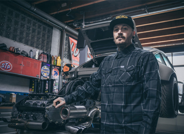 Man in a garage with a car and automotive products in the background - The Dapper Flannel by Greasy Hands Society