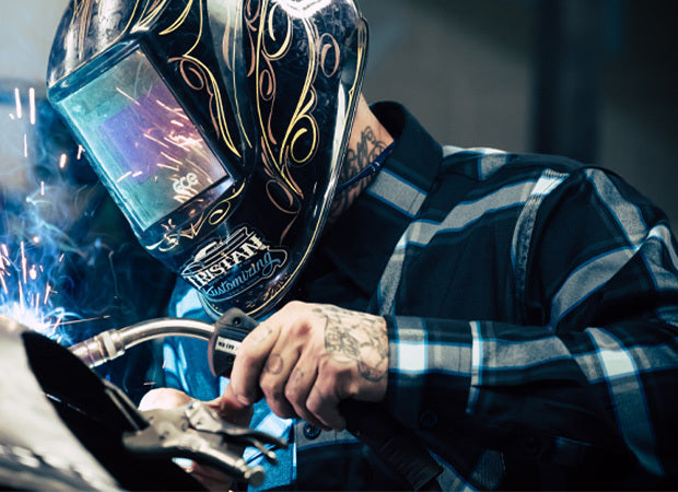 Person wearing a welding helmet with intricate designs, focusing on welding work - Gravity Fed Flannel by Greasy Hands Society