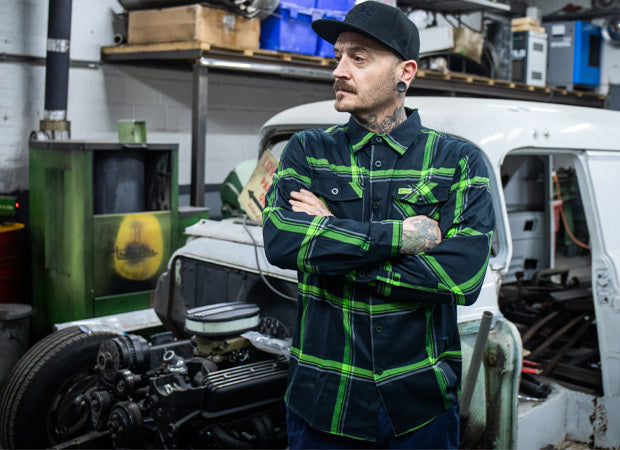 Man wearing a black and green plaid flannel shirt with green accents in a workshop setting - The Moto Flannel by Greasy Hands Society