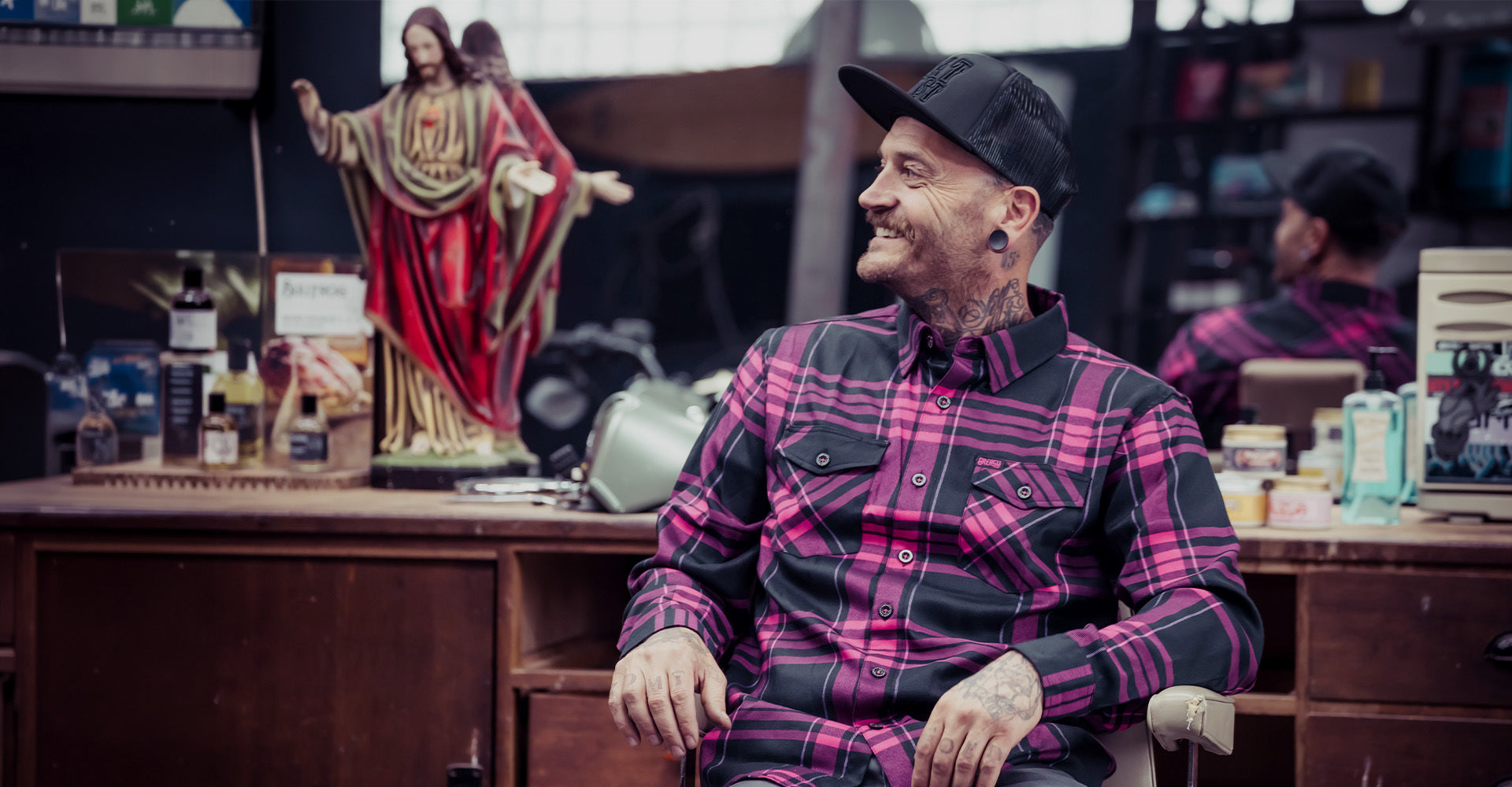 Man in a pink and black plaid shirt sitting in a barbershop with religious statue and various items on shelves - Greasy Hands Society