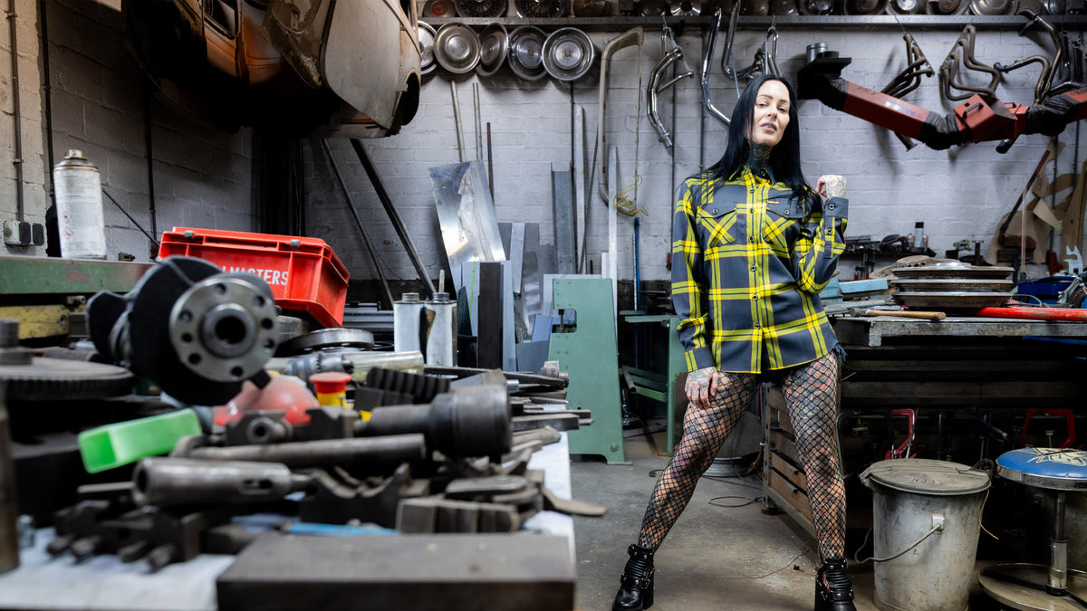A woman stands in a cluttered auto repair garage wearing a yellow and black plaid flannel shirt, fishnet tights, and black boots, surrounded by car parts and tools Women's mecum flannel in yellow and gray - Greasy Hands Society
