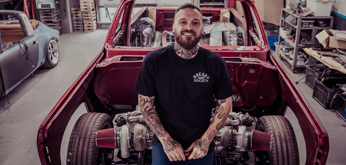 Man wearing a black greasy hands society t-shirt with tattoos standing in front of a red car with an open hood in a workshop.
