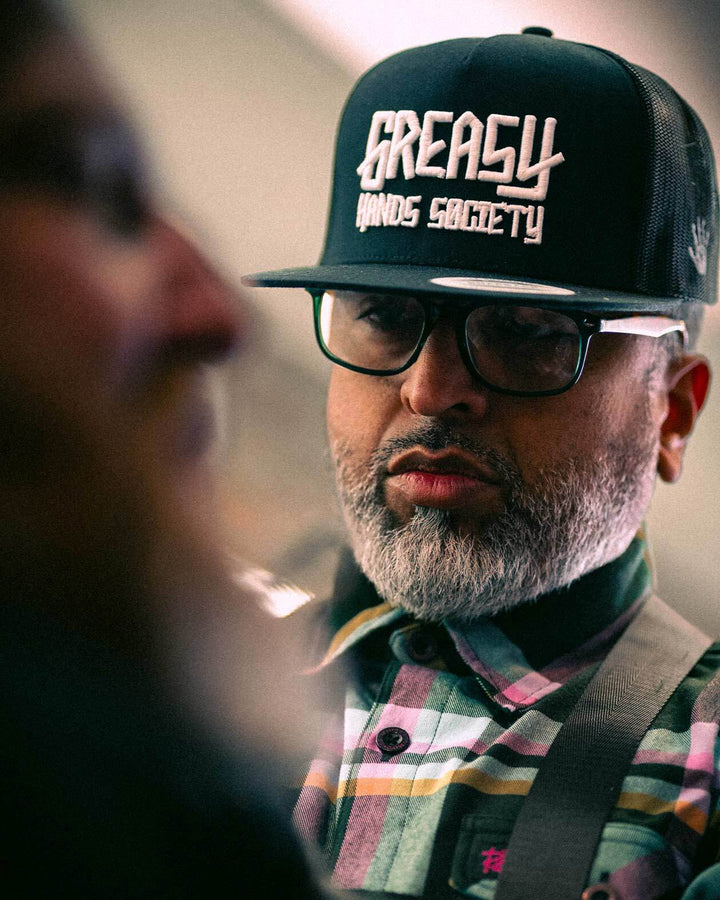Man wearing a 'Greasy Hands Society' cap with a blurred background - GHS Logo Black hat with white text by Greasy Hands Society