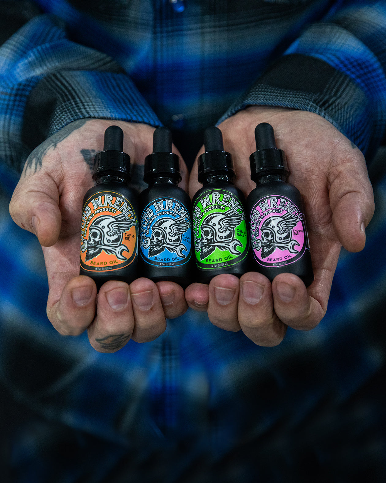 Four small bottles of psycho wrench beard oils held in hands against a blue plaid shirt background