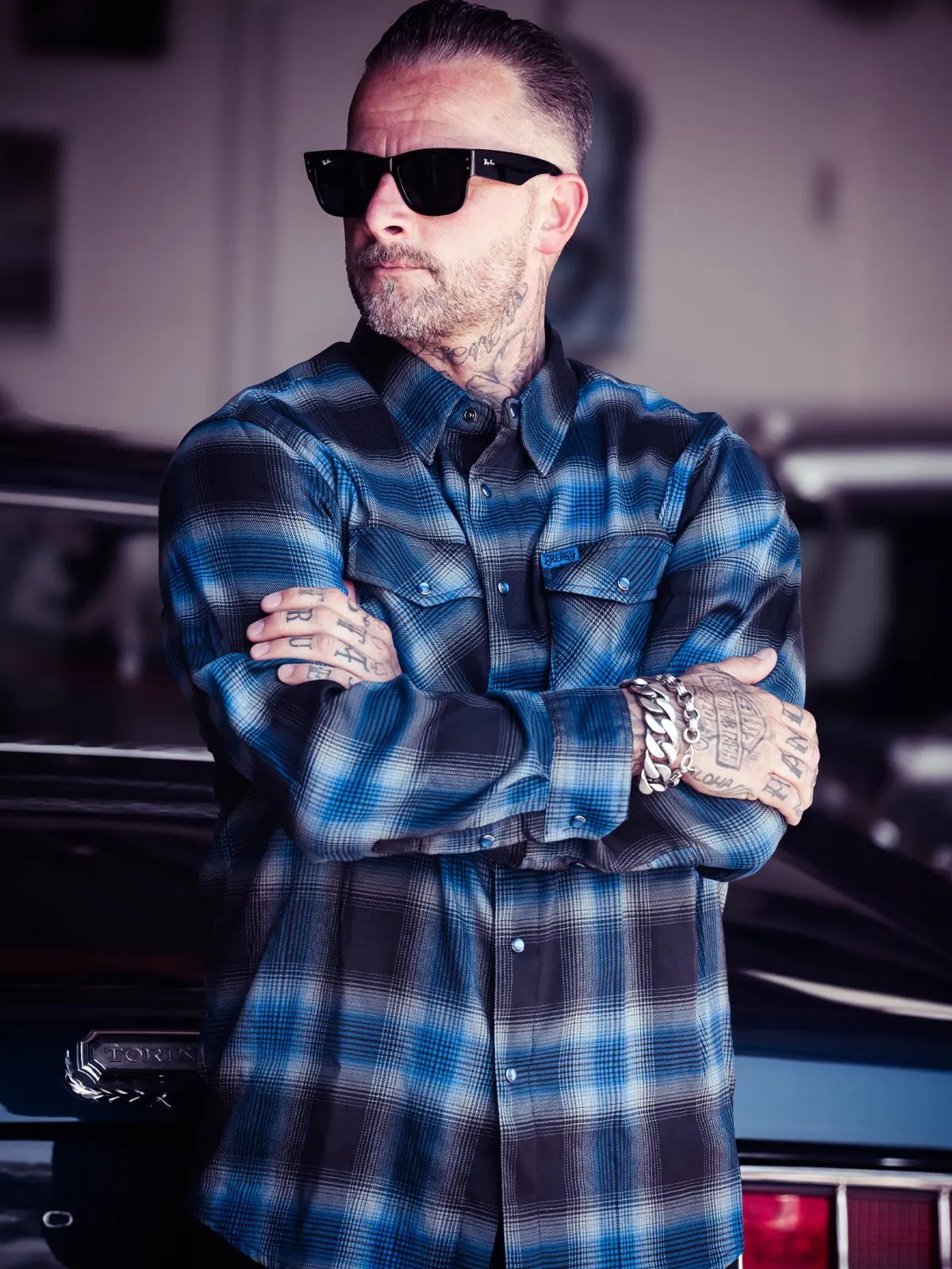A man looking cool wearing a black and blue plaid flannel shirt in a car workshop The Nailhead Flannel by Greasy Hands Society