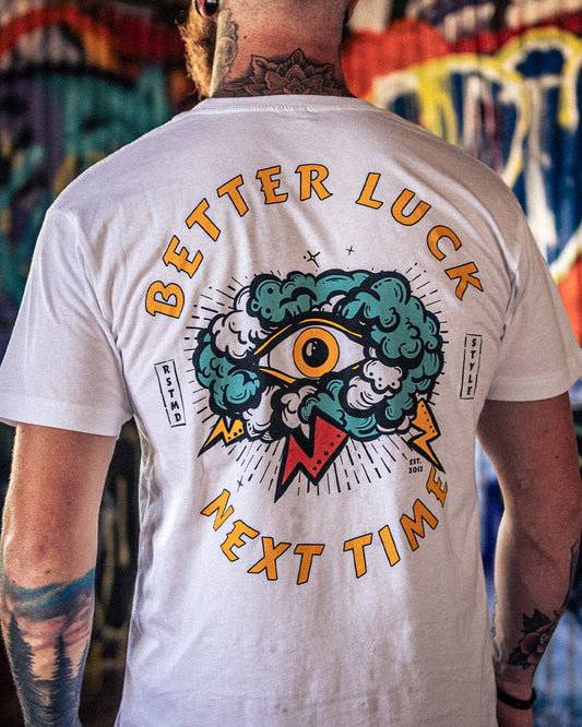 Person wearing a white t-shirt with a colorful graphic and text 'Better Luck Next Time' on the back, standing in front of a graffiti wall - Better Luck Next Time graphic t-shirt by Greasy Hands Society