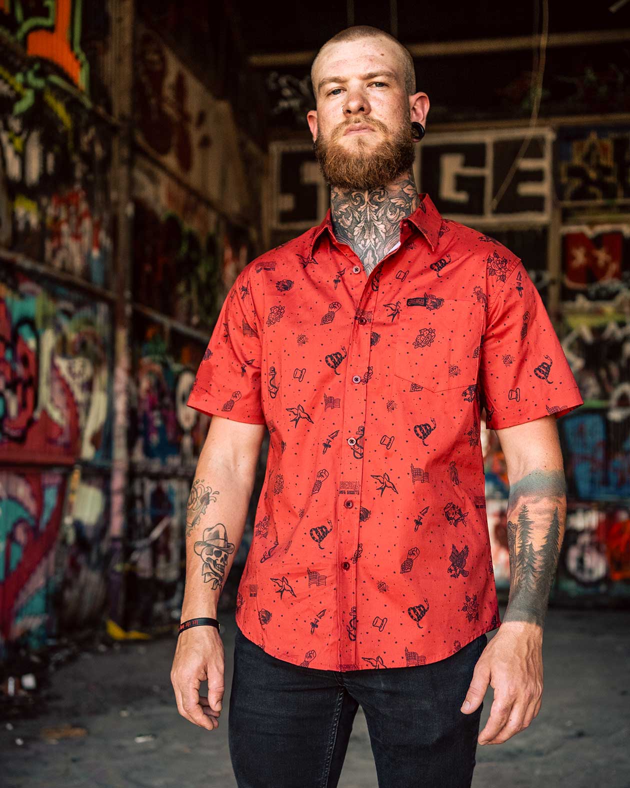 Man wearing a red patterned shirt standing in front of a colorful graffiti wall - The 1776 button-up short-sleeved shirt by Greasy Hands Society