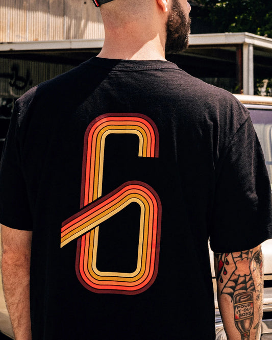 Person wearing a black t-shirt with a large orange and red number '6' on the back, standing outdoors - G70 graphic tee from Greasy Hands Society