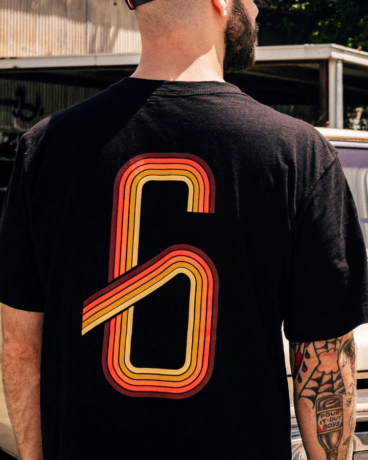 Person wearing a black t-shirt with a large orange and red number '6' on the back, standing outdoors - G70 graphic tee from Greasy Hands Society