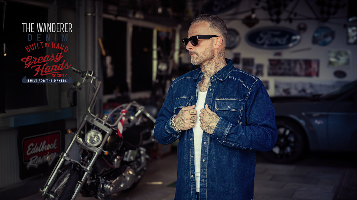 A man wearing a blue denim jacket over a white t-shirt stands in a garage-like setting with a motorcycle and a Ford-branded vehicle in the background. The text 'THE WANDERER DENIM BUILT BY HAND Greasy Hands' is visible on the left - The Wanderer Denim Shirt by Greasy Hands Society