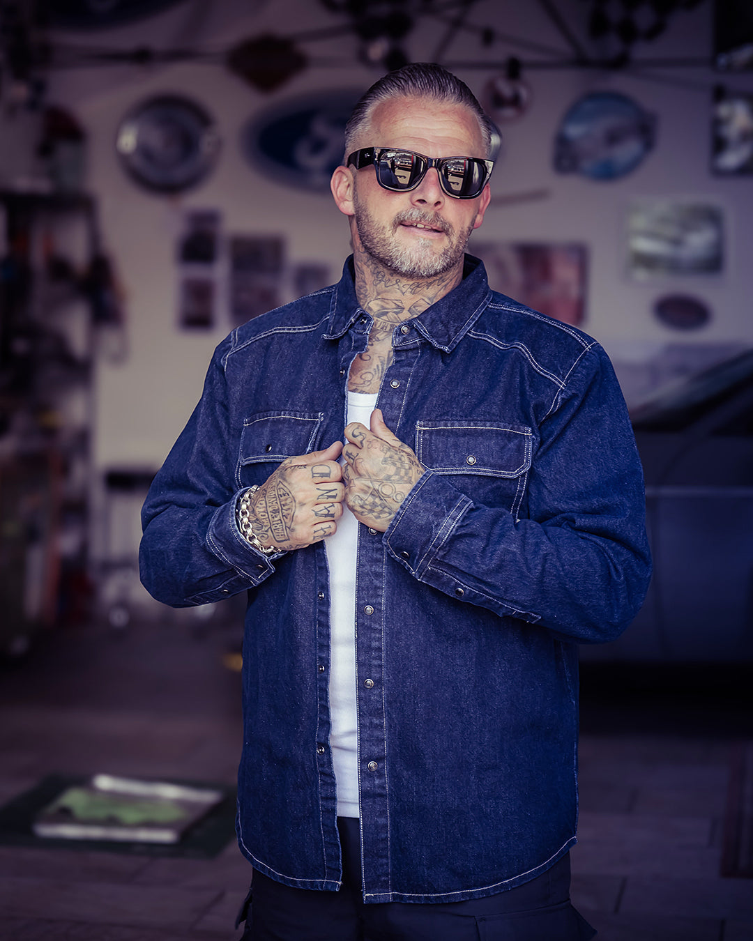 A man wearing a dark blue denim jacket over a white t-shirt, styled with black sunglasses and bracelets, standing in a garage-like environment - The Wanderer Denim Shirt by Greasy Hands Society