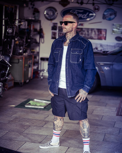 A man wearing a dark blue denim button-up shirt, dark cargo shorts, a white t-shirt, and checkered slip-on shoes stands in a garage-like workshop environment - The Wanderer Denim Shirt by Greasy Hands Society